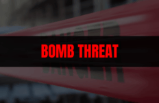 News graphic with a blurred background and red-and-white police tape, overlaid with a black banner reading ‘BOMB THREAT’ in bold red letters.
