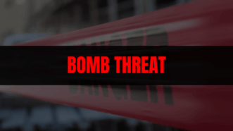 News graphic with a blurred background and red-and-white police tape, overlaid with a black banner reading ‘BOMB THREAT’ in bold red letters.