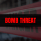 News graphic with a blurred background and red-and-white police tape, overlaid with a black banner reading ‘BOMB THREAT’ in bold red letters.