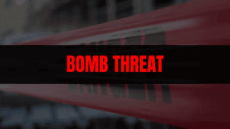 News graphic with a blurred background and red-and-white police tape, overlaid with a black banner reading ‘BOMB THREAT’ in bold red letters.