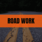 A two-lane road with double yellow lines, with an orange 'ROAD WORK' banner across the center.